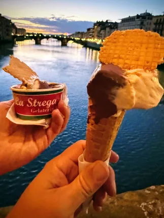 Two hands holding gelato, a cone and a La Strega tub, above the Arno at sunset with a lit bridge behind