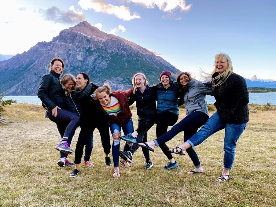 Eight women and girls posed together laughing in front of a mountain and blue glacier lake, arms linked