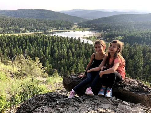 The two girls sitting together on a rock high above a forested valley and lake at Whitefish