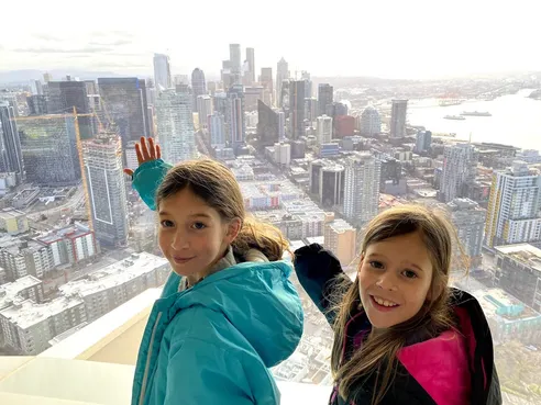 Two of the girls at the top of the Space Needle, one waving, downtown Seattle and the waterfront far below
