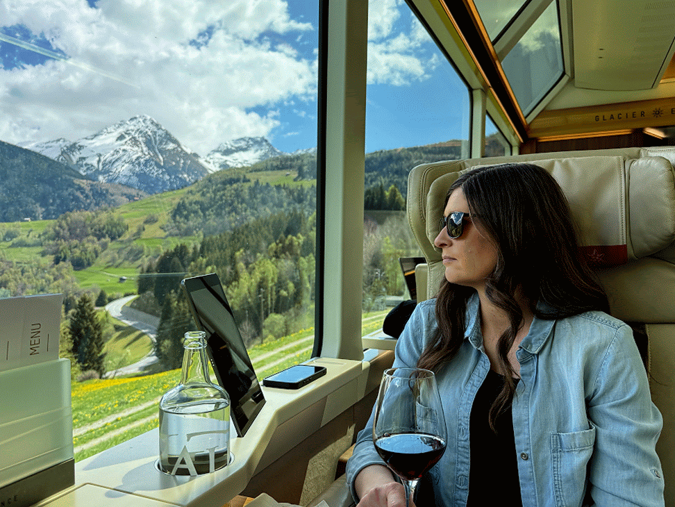 Emily in her armchair by the Glacier Express window with a glass of red wine, the snowy peak and green valley sliding past outside