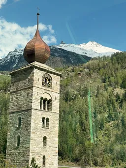 A stone bell tower with a copper onion dome and a clock face, a snow-covered peak rising behind the wooded slope