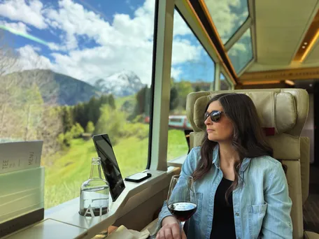Emily in a leather armchair by the panoramic window of the Glacier Express with a glass of red wine, green pasture and a snowy peak outside