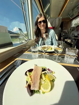 A plated fillet of fish on salad with lemon, wine glasses set on the table and Emily smiling across it