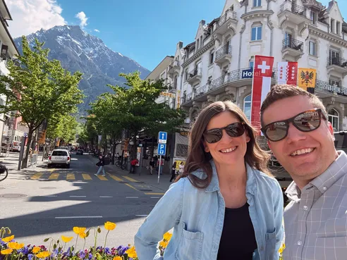 Emily and Josh on a town street lined with trees and hotels, a snow-topped peak at the end of it and tulips in the foreground