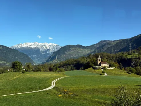 A small stone church on a green knoll with a track winding up to it, a snow-covered range across the valley