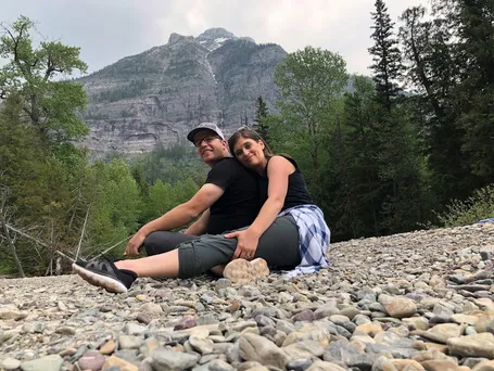 Emily leaning against Josh on a bed of river stones, a steep grey mountain face rising behind the treeline