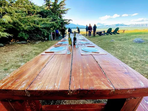A long wooden table set for dinner in a field with mountains and a glacial lake behind, people standing by