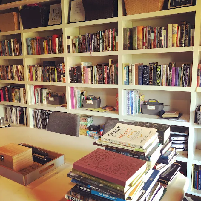 The home library wall of white cube shelves packed with books, a desk in front stacked with more