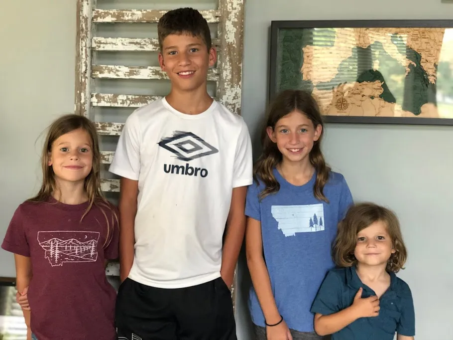 The four children lined up by height in front of an old shutter and a wooden map, the youngest at the end