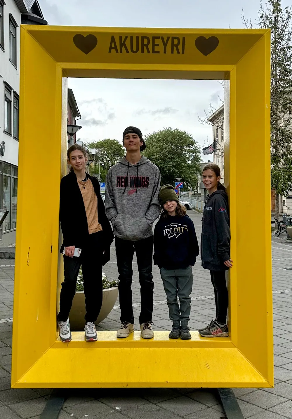 The four children standing inside a big yellow picture frame on the street that reads Akureyri between two hearts