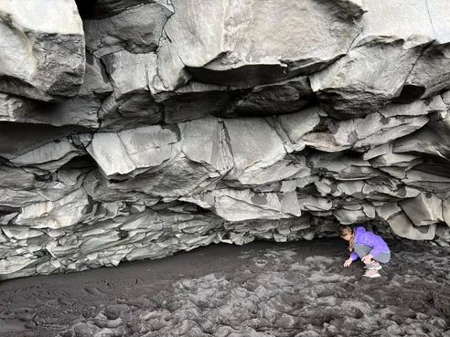 Mia in a purple jacket crouching under the low overhang of stacked basalt slabs on the black sand