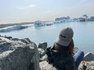 Case in a cap seen from behind, sitting on the rocks looking out over the icebergs in the glacier lagoon