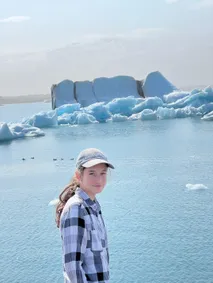 Evie in a checked shirt and cap turning to the camera, a big blue iceberg on the pale lagoon behind her