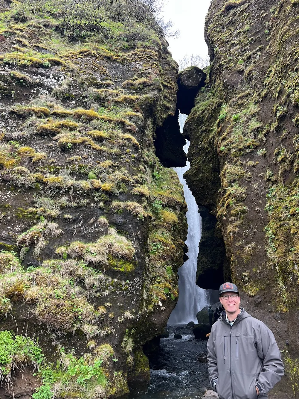 Josh in a grey jacket and cap at the mouth of the narrow mossy cleft that hides Gljufrabui, the waterfall falling through the gap behind him