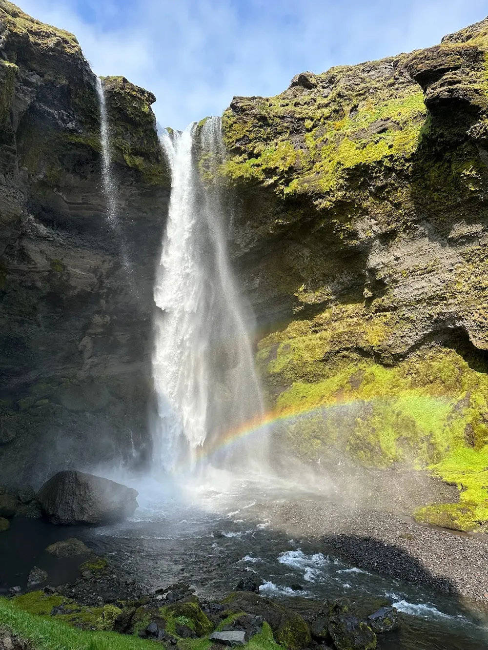 Kvernufoss falling into a rocky gorge with a bright rainbow thrown across the mossy wall beside it