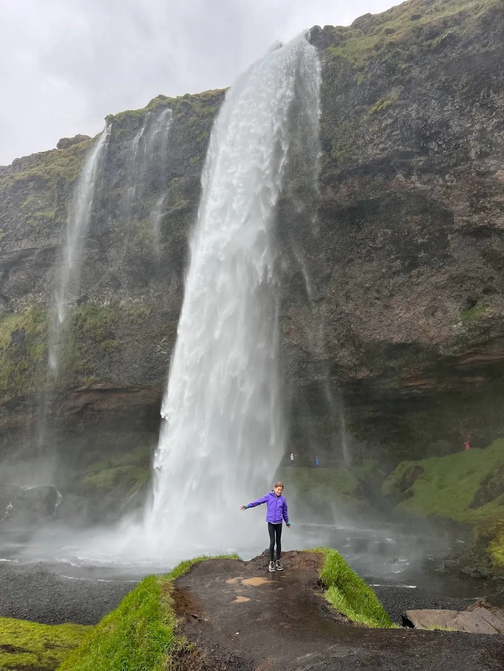 Mia in a purple jacket with her arms out on the wet rock below Seljalandsfoss, the fall dropping from the mossy cliff behind her