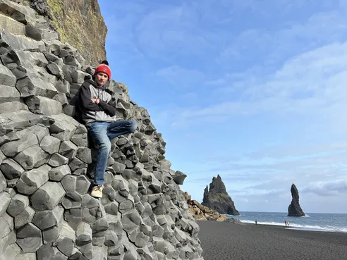 Preston in a red bobble hat leaning back against the hexagonal basalt columns, the black beach and sea stacks behind him