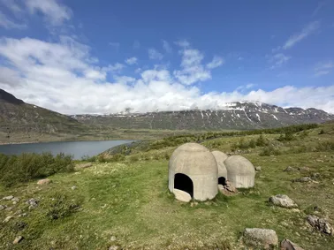 Three concrete acoustic domes standing on the grass above the fjord, cloud sitting along the snow-streaked ridge opposite