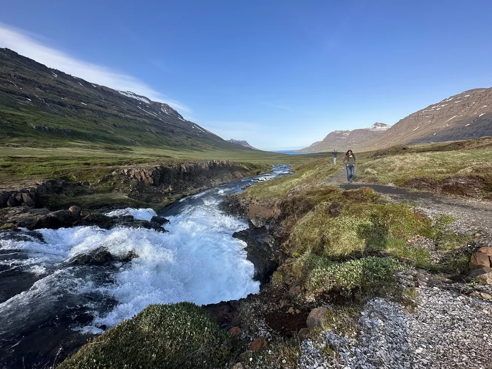 White water pouring over a rock lip into a valley that runs down to the fjord, Emily walking the path on the right