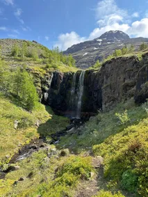 A waterfall dropping over a dark rock step into a green valley, a snow-patched peak rising behind