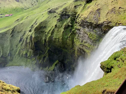 Skogafoss seen from the clifftop steps, the water dropping into a dark pool with a red-roofed farm on the green flats beyond