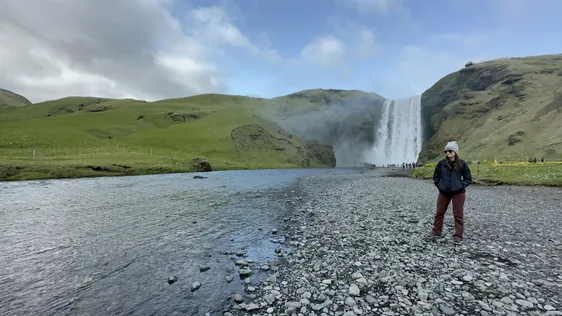 Emily in a knit hat on the river stones below Skogafoss, the fall dropping between green hillsides behind her