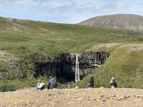 Four of them sitting on the gravel rim watching Svartifoss drop over its wall of dark hanging basalt columns