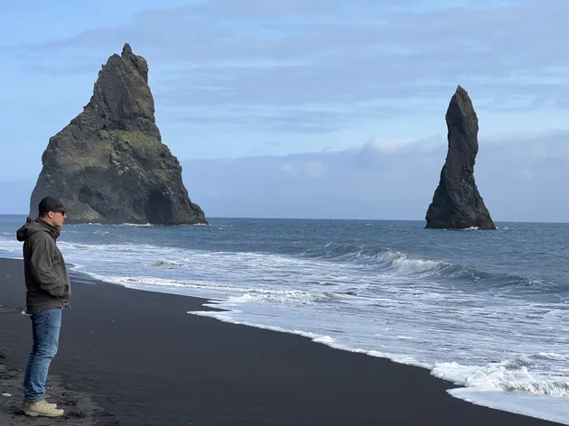 Josh at the tide line on the black sand at Reynisfjara, the two Reynisdrangar sea stacks standing out of the surf