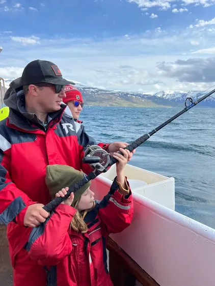 Josh helping the youngest hold a heavy fishing rod over the side of the boat, another of the children watching, snow-topped mountains across the fjord