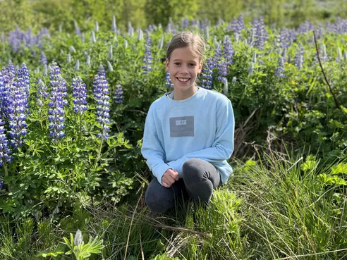 Mia crouching and grinning in a field of purple lupins in a pale blue sweatshirt