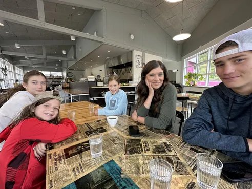 The five of them round a cafe table with newspaper print under the glass, mugs and water glasses in front of them, the open kitchen behind