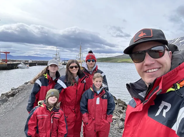 All six in red and navy flotation suits on the quay before the whale watching trip, boats and the fjord behind them