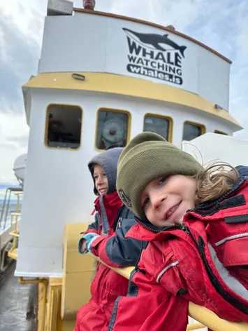 Two of the children in red flotation suits leaning on the rail of the whale watching boat below its wheelhouse