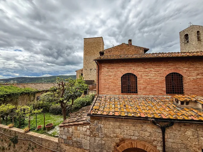 The brick flank and bell tower of a church in San Gimignano above lichened roof tiles, a stone tower and the hills beyond