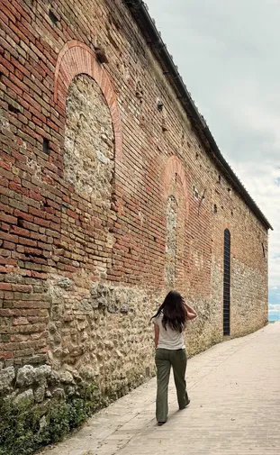 Emily walking a paved lane beside a long brick and stone wall with blocked arches