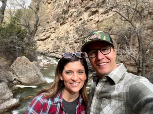 Josh and Emily close together in front of a creek running below a limestone cliff