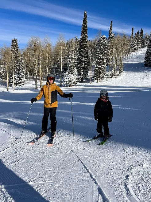 Josh in a yellow jacket and Case on their skis on a groomed run, snow-laden pines behind them