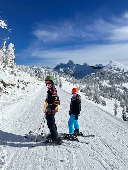 Preston and Mia on their skis at the top of the run in helmets and goggles, frosted firs and the Teton peaks behind