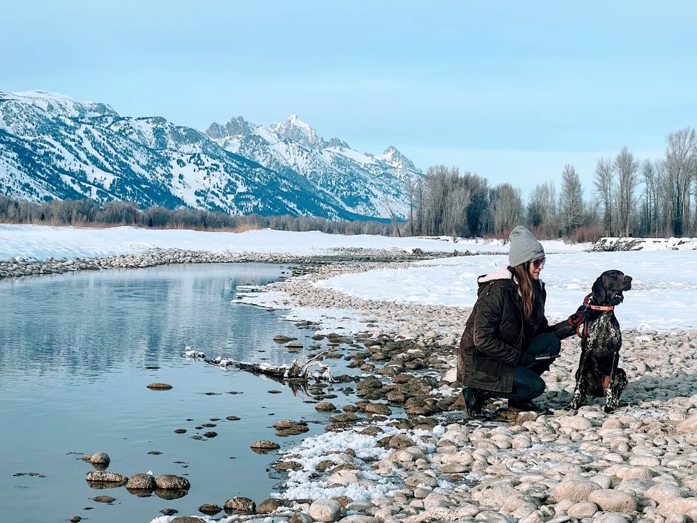 Emily in a knit hat crouching on the snowy stones of a river bank beside the dog, the Tetons rising white behind them