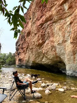 Josh sitting in a camp chair in the creek shallows beneath a great overhanging red and white cliff, the dog wading past