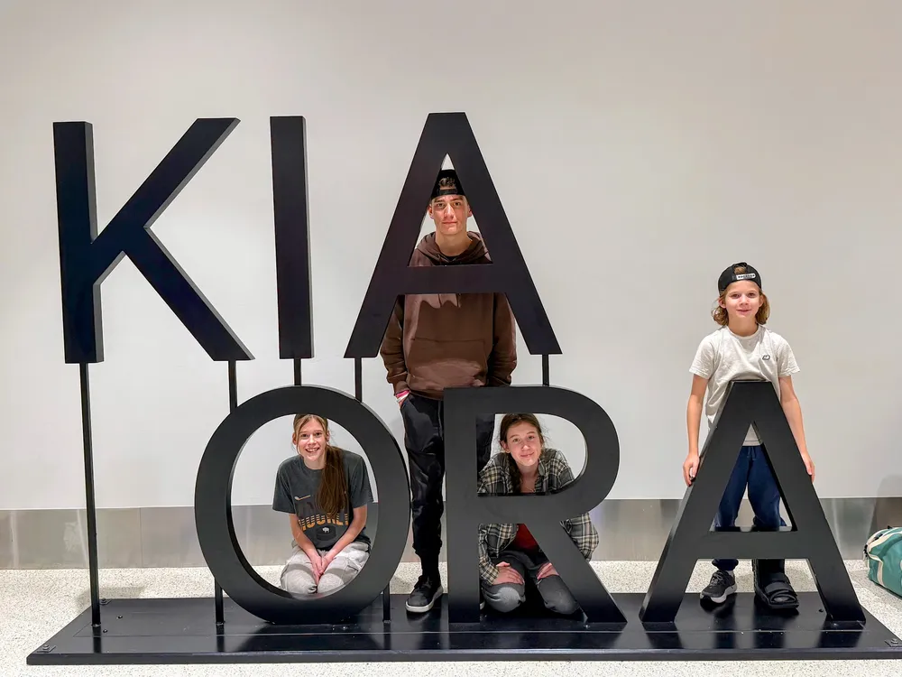 The four children arranged in the big black KIA ORA letters at the airport, one sitting inside the O, Preston standing behind the A, another kneeling in the R, and Case at the end in a walking boot