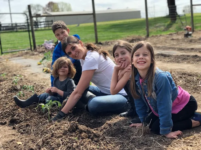 Emily kneeling in the garden bed with all four children around her, seedlings just planted in the turned soil