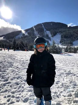 Case in a black ski jacket, helmet and orange goggles standing on the snow, the mountain runs behind him
