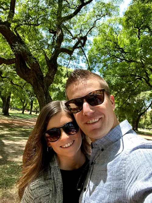 Emily and Josh together smiling in the sun on a park bench under tall shade trees
