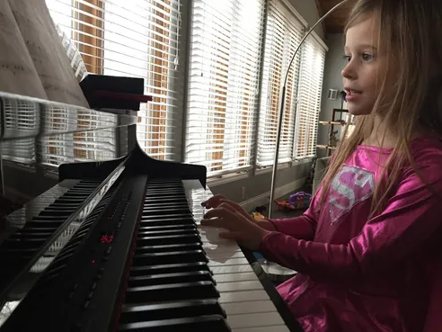 One of the girls in a pink shirt playing the keyboard by a window of venetian blinds, sheet music propped in front of her