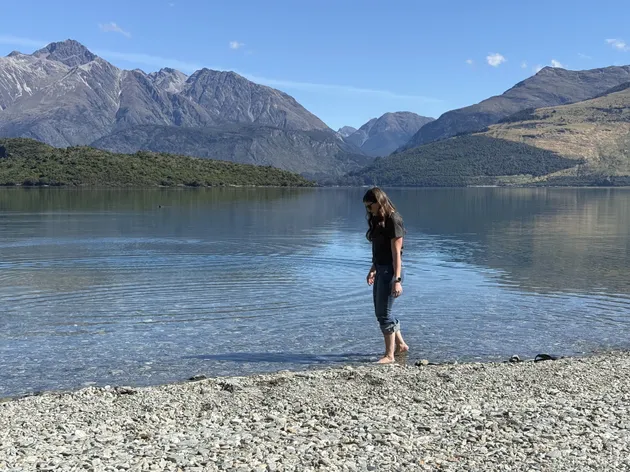 Emily standing barefoot in the clear shallows of the lake at Glenorchy, jagged peaks reflected in the still water behind her