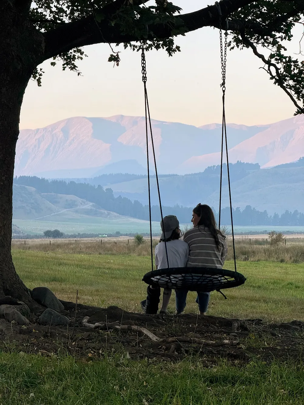 Emily and Case sitting together on a round rope swing hung from a big tree, looking out at pink lit mountains across the fields at dusk