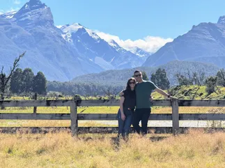 Emily and Josh leaning on a post and rail fence in the long grass, a glacier and snow-covered peaks filling the view behind