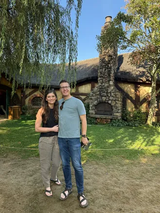 Emily and Josh together on the lawn outside the Green Dragon, the thatched roof and stone chimney behind them and a willow overhead
